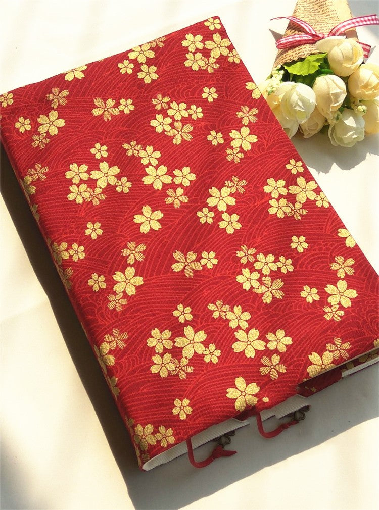 Red fabric book cover with a gold-tone cherry blossom and wave print on a book, beside white flowers