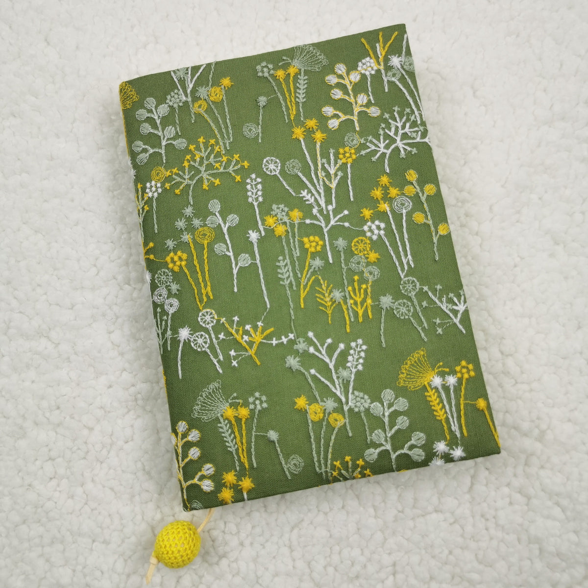 Green fabric book jacket with embroidery-style yellow and white wildflowers and a cord with a yellow crocheted bead
