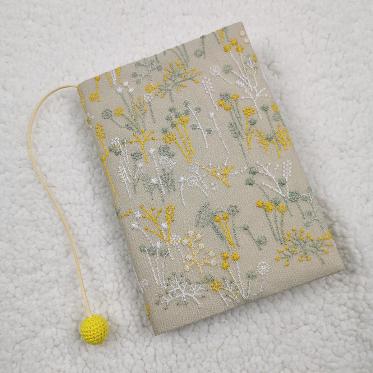 Beige fabric book jacket with embroidery-style yellow and green wildflowers and a cord with a yellow crocheted bead