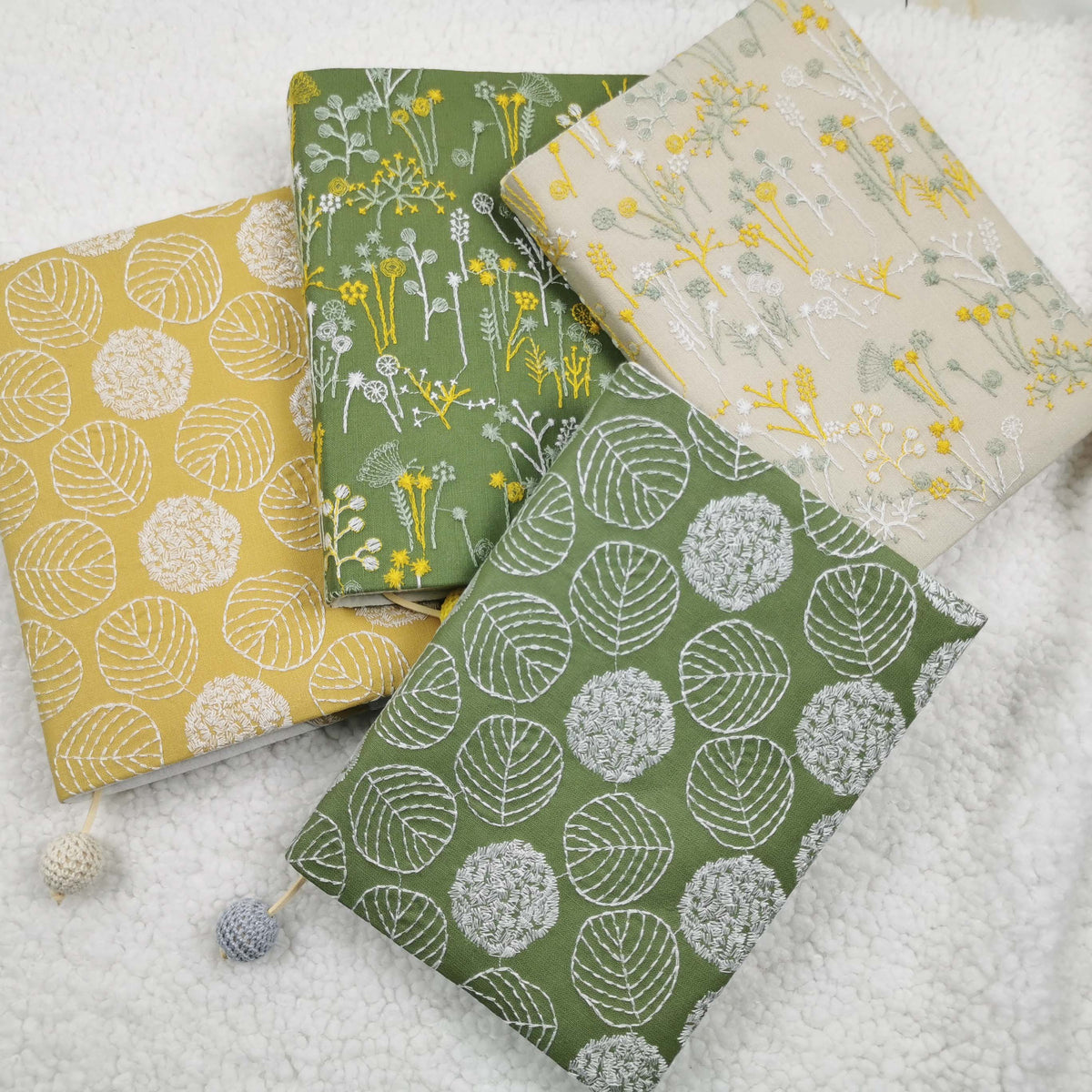 Four embroidery-style fabric book jackets in yellow, green and beige designs with crocheted bead cords