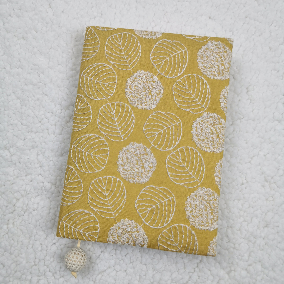 Yellow fabric book jacket with white embroidery-style leaf outlines and a cord with a cream crocheted bead