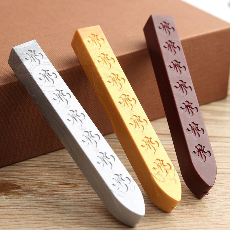 Three sealing wax sticks in silver, gold and burgundy with an embossed pattern, lying against a kraft box